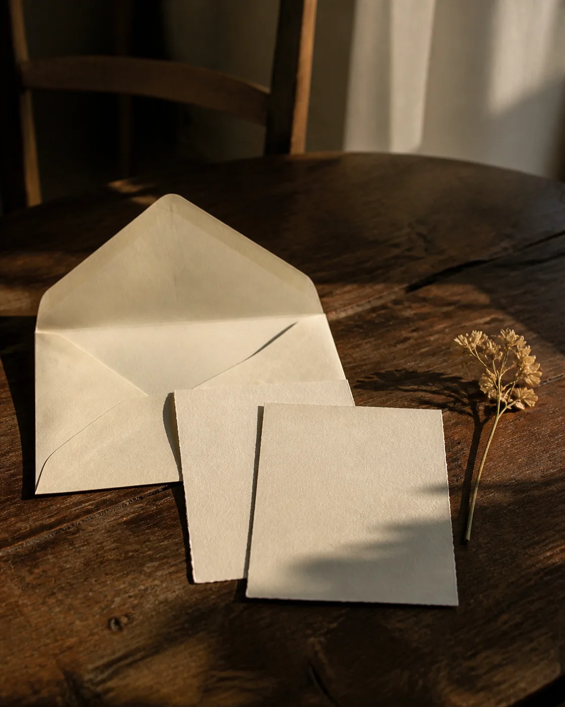 An open cream envelope, blank cards, and a dried flower arranged on a worn wooden table