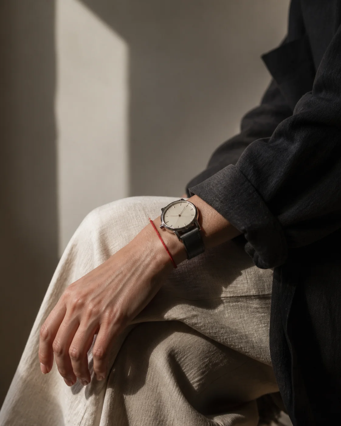 An anonymous wrist wearing a fine red thread beside a simple watch on dark linen