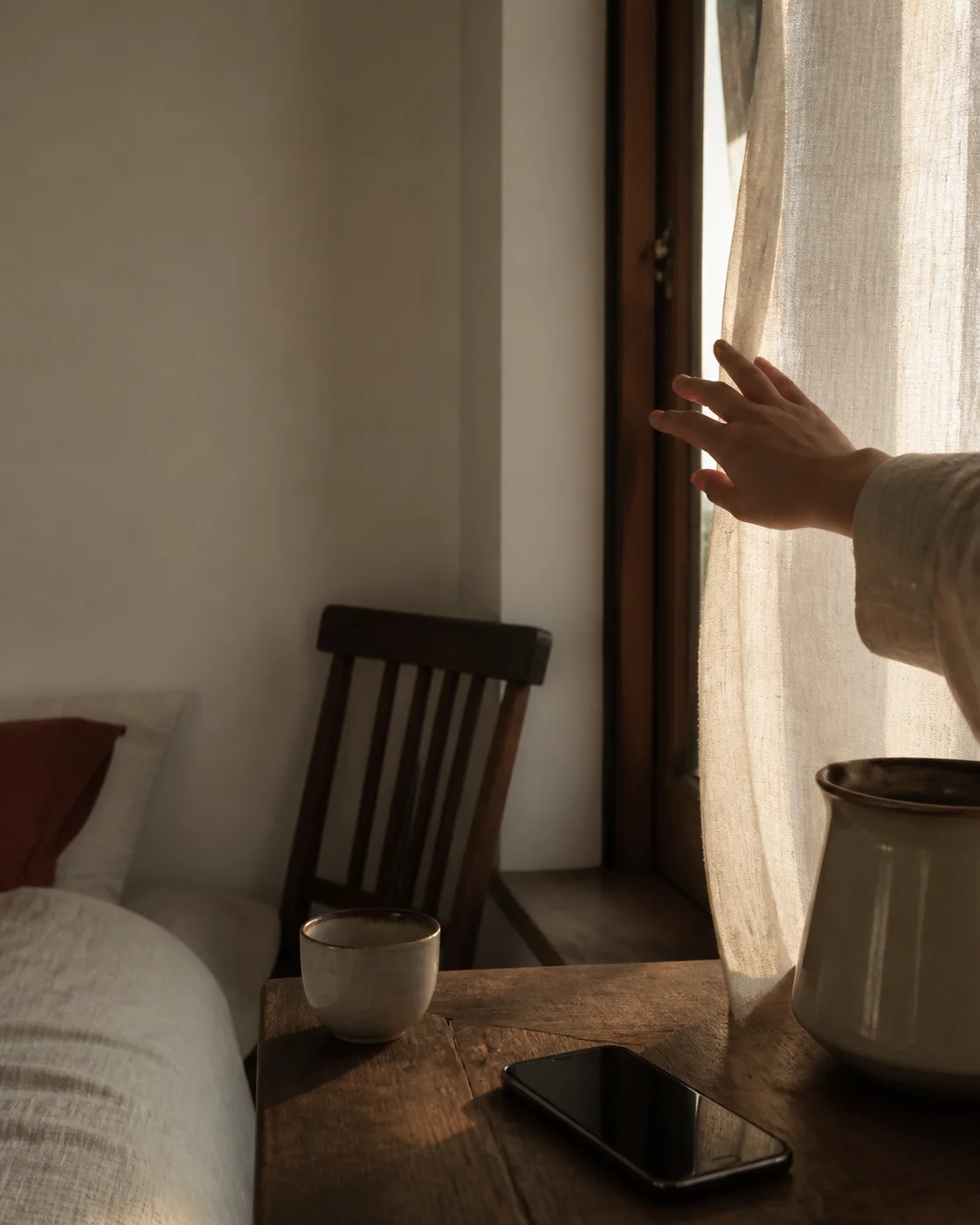 A hand opening a linen curtain beside a ceramic cup before the day begins